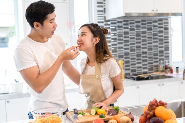 A young Asian man is feeding his wife a piece of sliced tomato in the kitchen.