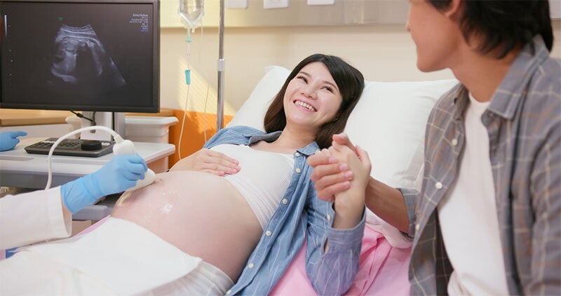 A young Asian woman holding her husband’s hand and smiling while lying in a hospital bed as a doctor performs an ultrasound scan.