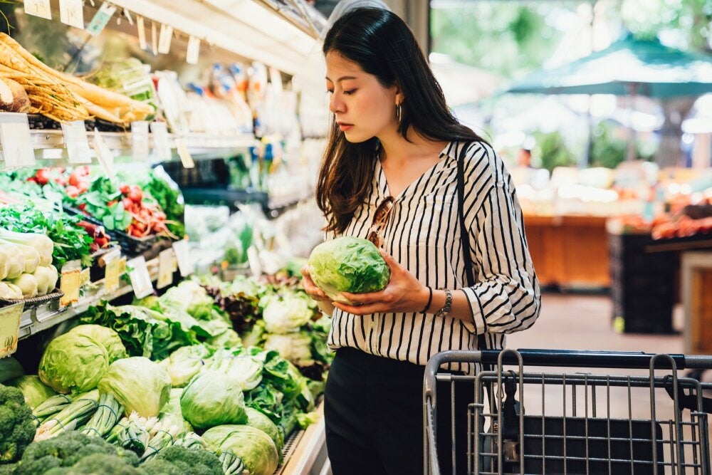 Asian female in stripes looking at various leafy greens in grocery store.