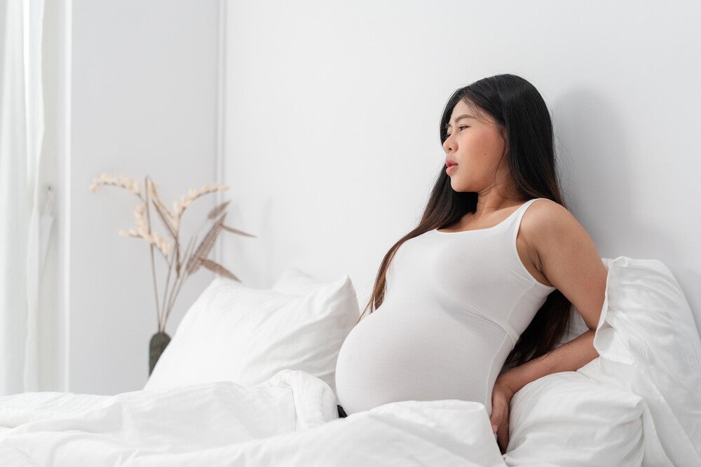 Pregnant Asian woman in white holding lower back as if in pain.