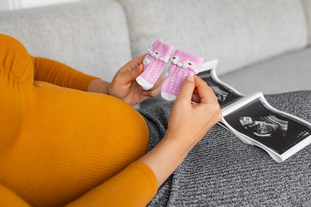 Female hands hold a pair of pink socks near baby bump with ultrasound pictures on her lap.