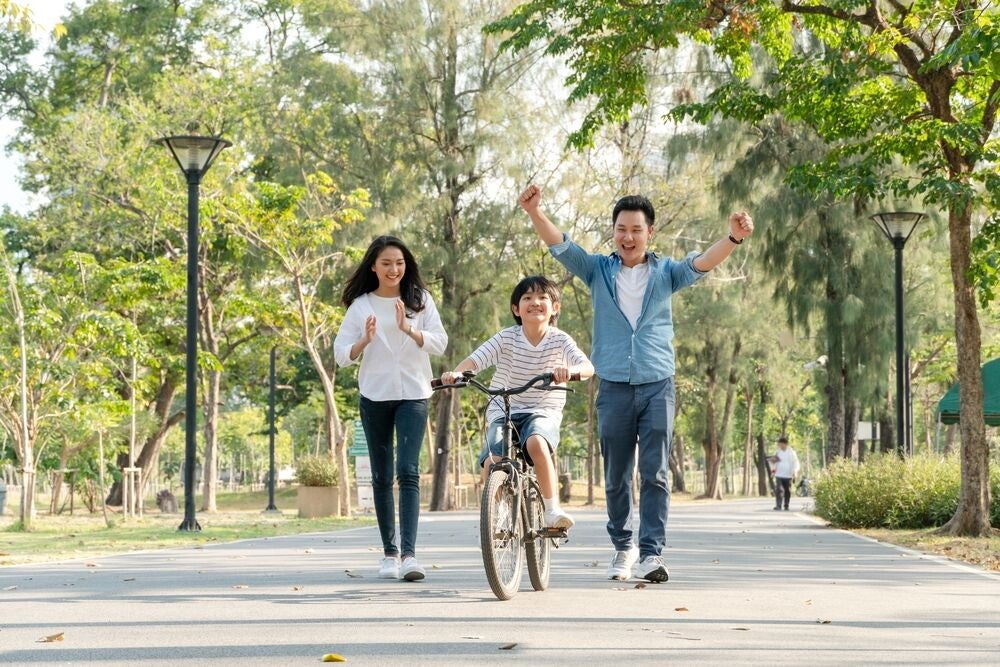 Asian parents cheer on their son as he bikes on his own at a park.
