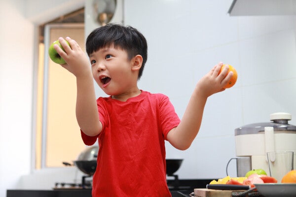 A little Asian boy holds an apple and an orange in each hand in the kitchen.
