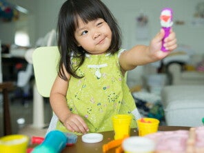 Teaser_Image Asian preschool-age girl lifts a toy and tilts her head as she looks at it with one eye closed.