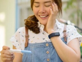 Asian woman in denim jumping smiling at pregnancy test results.