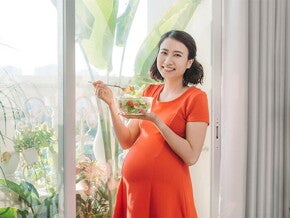 A young Asian woman standing by the window and holding a bowl of salad as part of her healthy pregnancy meal plan A young Asian woman standing by the window and holding a bowl of salad as part of her healthy pregnancy meal plan