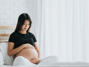 A young pregnant Asian woman holding her belly and seated in bed, alert to the signs of labor A young pregnant Asian woman holding her belly and seated in bed, alert to the signs of labor