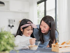 Asian mother watching her child eating cereal from a bowl Asian mother watching her child eating cereal from a bowl