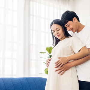 A 26 weeks pregnant woman holding her belly while her husband hugs her from behind in their home A 26 weeks pregnant woman holding her belly while her husband hugs her from behind in their home