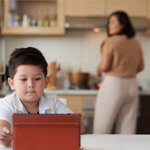 A little Asian boy watching a show on a tablet while his mother is cooking dinner in the background