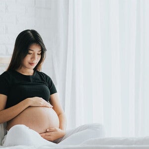 A young pregnant Asian woman holding her belly and seated in bed, alert to the signs of labor