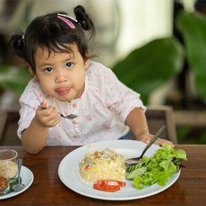 Cute Asian little girl eats fried rice with tomatoes and green leafy vegetables on the side