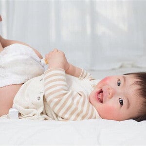 Hands holding a smiling Asian baby’s legs and removing the diaper to check baby poop color. 