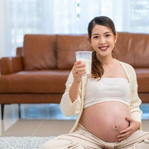 Pregnant Asian woman holding a glass of milk in front of brown sofa.