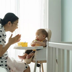 Young Asian mom claps her hands as she watches her baby eating in a high chair.
